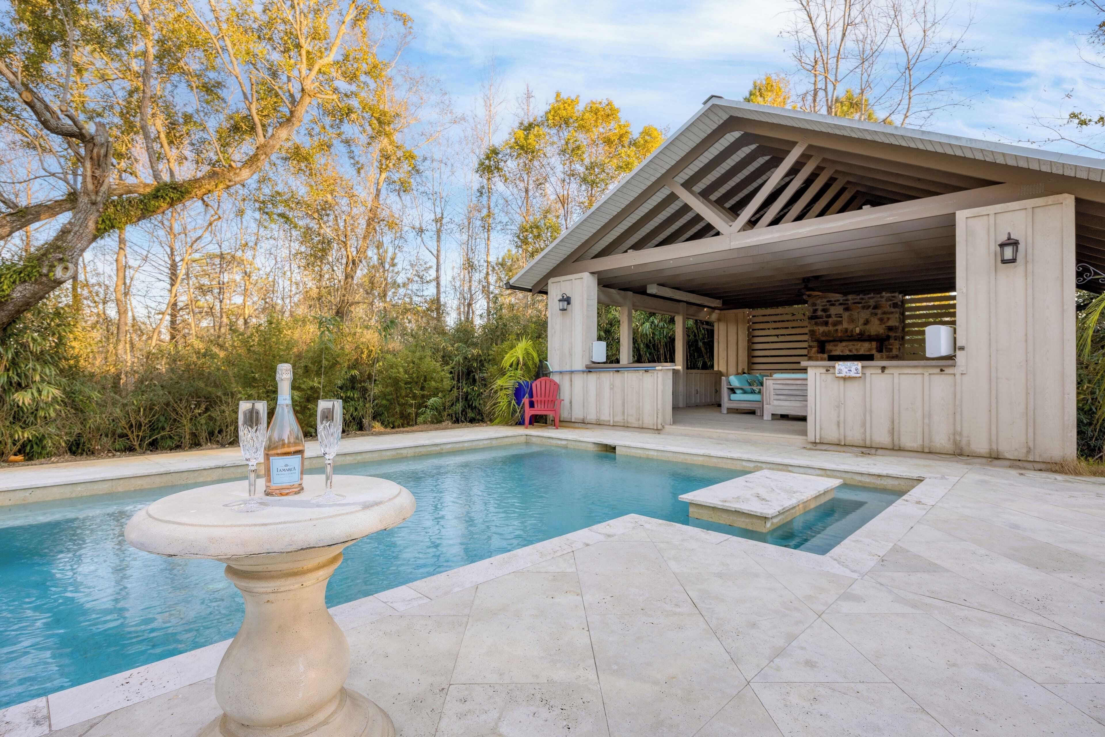 A pool area with a covered patio, a stone wall, and a table with a bottle and glasses.