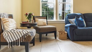 A living room with two armchairs, a small table with a vase of flowers, a blue sofa, and a window with a view of trees.