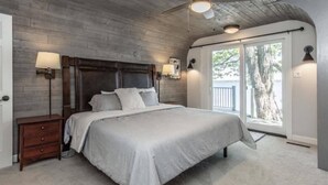 Une chambre avec un plafond en bois, un lit recouvert d’une couverture grise, deux lampes de chevet et une vue sur l’extérieur par une fenêtre.