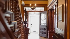 A grand hallway with a wooden staircase, a chandelier, and a door leading to a bright room.