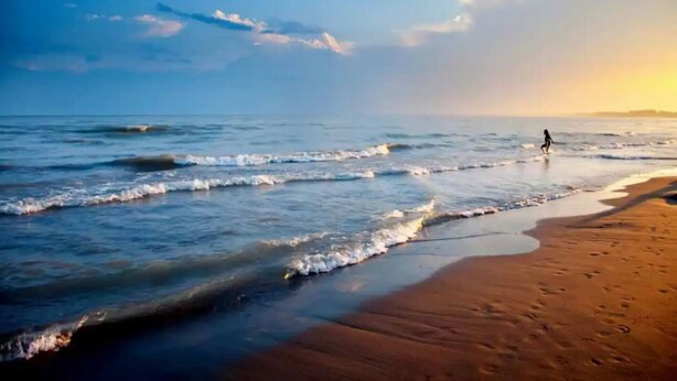 A beach with waves, a person walking, and a sunset.