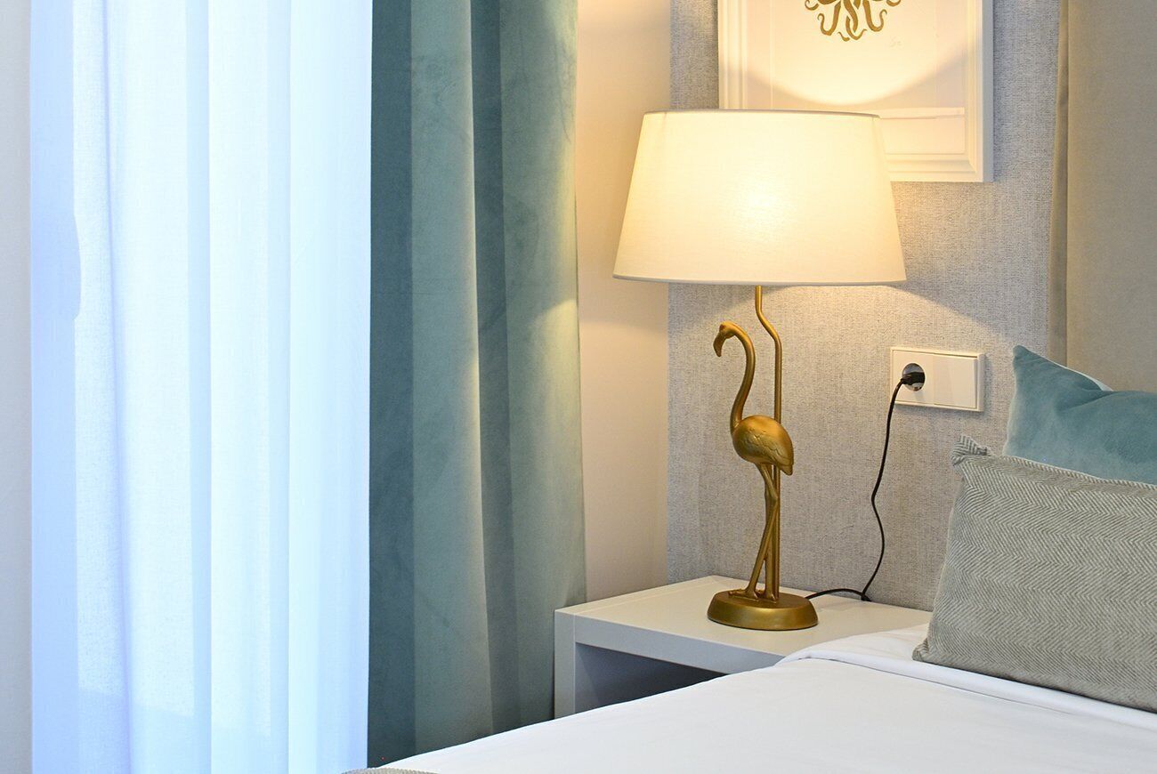 Een net opgemaakt bed met een witte deken en een lichtblauwe kussen. Een gouden lamp met een uniek ontwerp staat op een witte nachtkastje naast het bed. De kamer heeft een lichtblauwe gordijn en een muur met een lijstwerk.