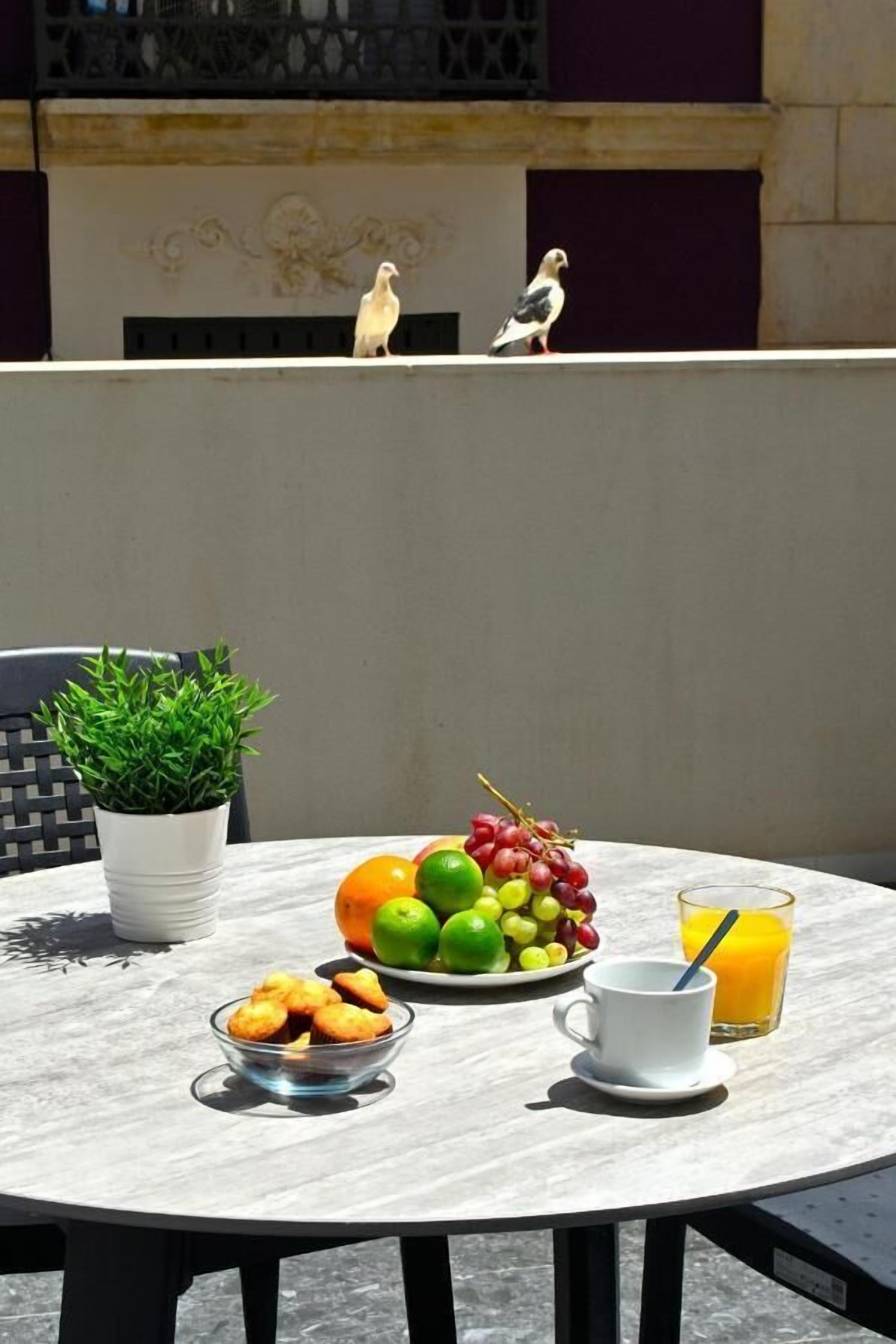 Una mesa redonda con un frutero, un vaso de jugo de naranja y una taza con cuchara. Una planta en maceta está sobre la mesa, y un pájaro está posado en la pared de fondo.