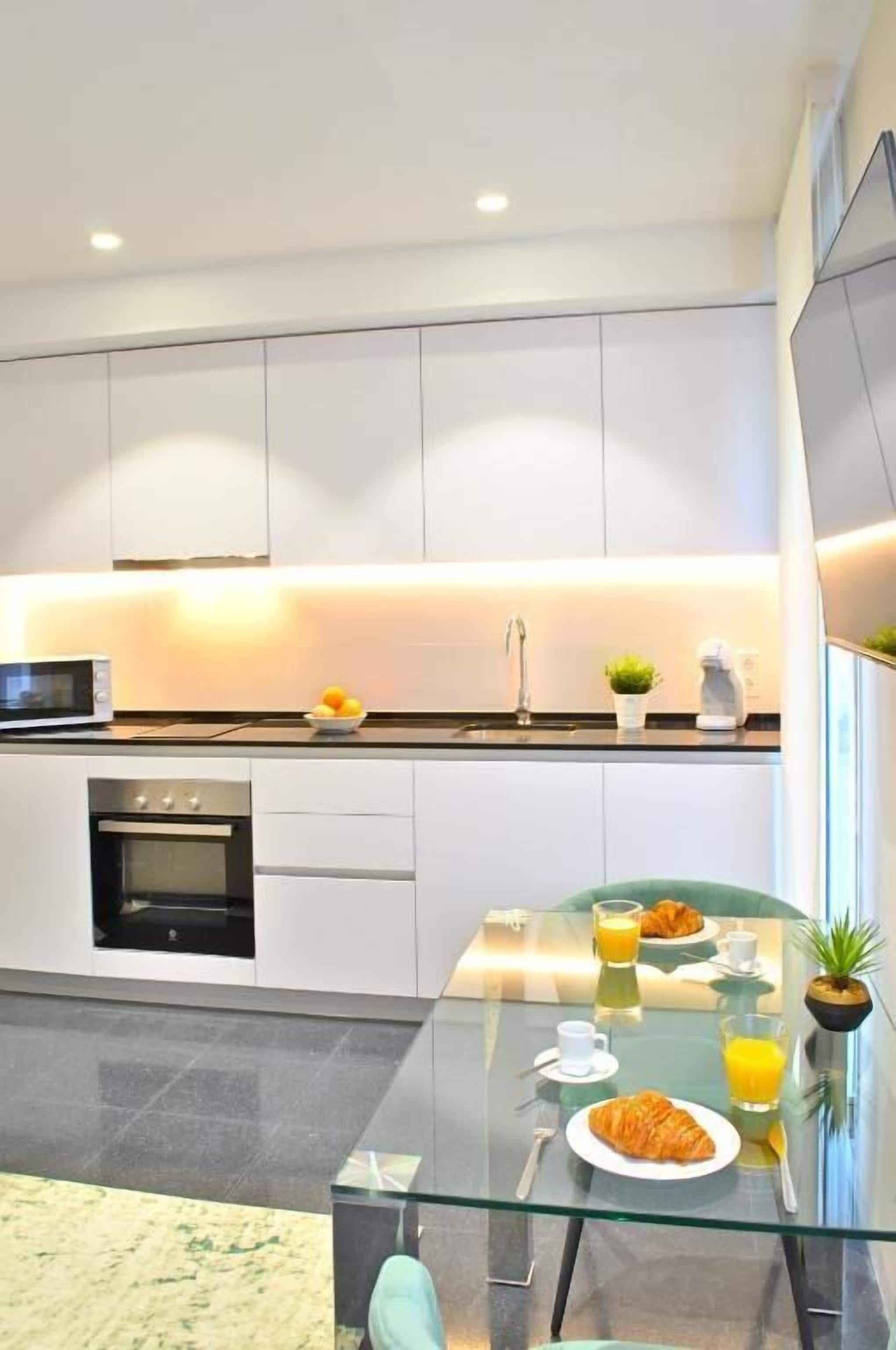 A modern kitchen with white cabinets, a glass dining table, and breakfast items.