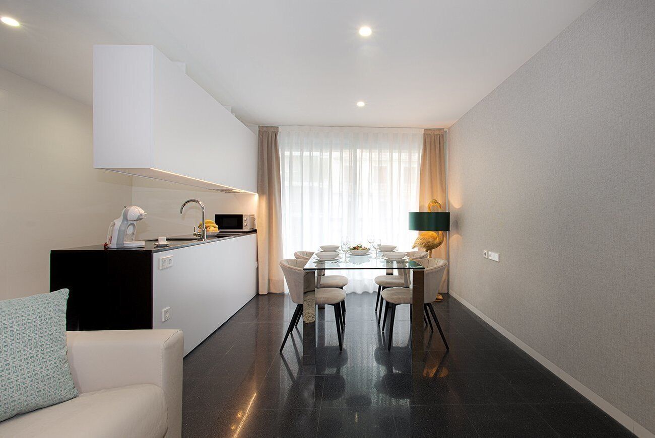 Une cuisine moderne avec un coin repas, comprenant une table et des chaises en verre, un plan de travail noir et un canapé blanc agrémenté d’un coussin à motifs.