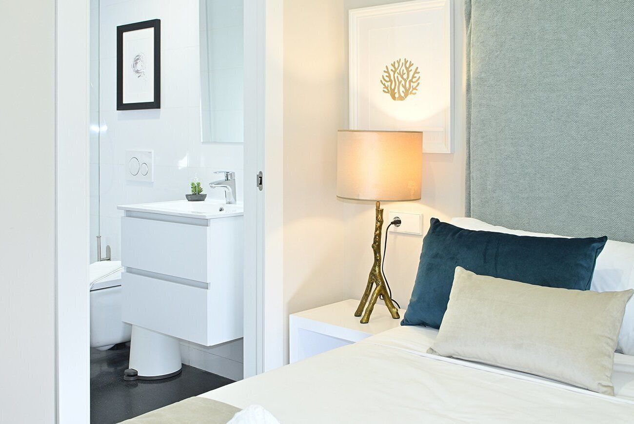 A modern hotel room with a white bed, a bedside table with a lamp, a framed picture on the wall, and a bathroom with a white sink and toilet.
