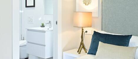 A modern hotel room with a white bed, a bedside table with a lamp, a framed picture on the wall, and a bathroom with a white sink and toilet.