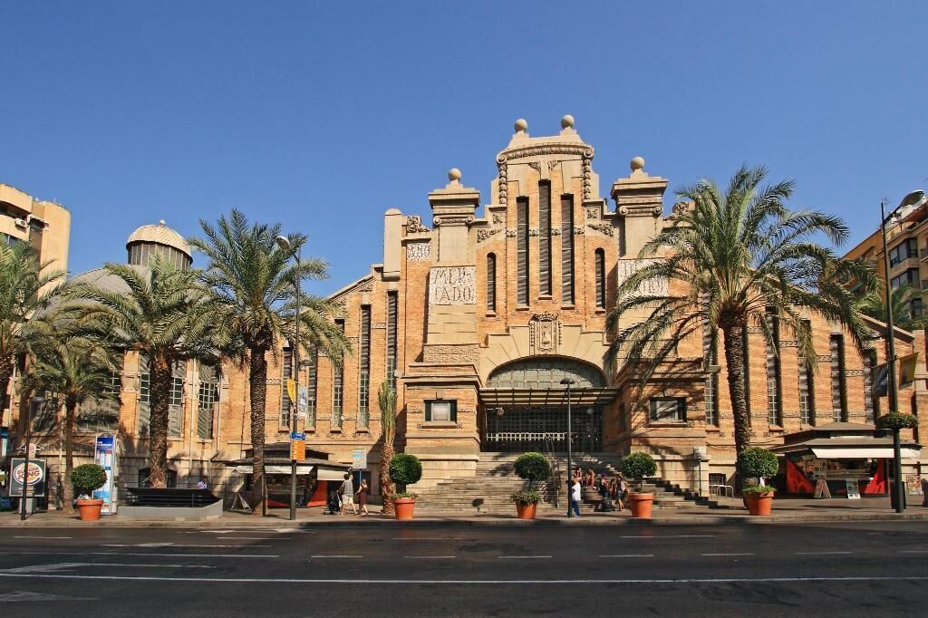 Un edificio histórico con una cúpula central y una entrada abovedada, rodeado de palmeras y plantas en macetas.