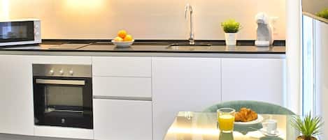 A modern kitchen with a built-in oven, a microwave, and a sink. There is a dining table with a glass top, a green chair, and a small potted plant.