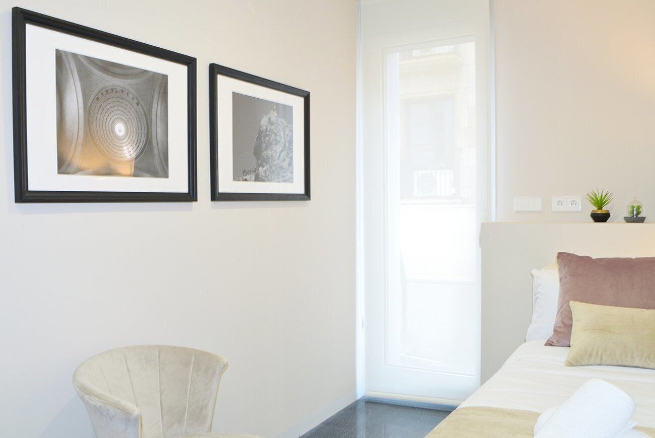 Habitación de hotel con una cama, una silla, dos cuadros en la pared, una ventana y una planta pequeña en la mesita de noche.