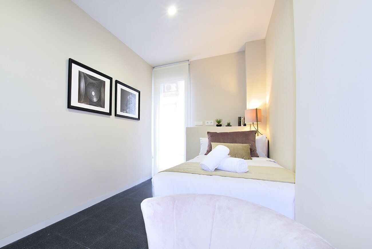 A hotel room with a bed, a chair, a desk with a lamp, and two framed pictures on the wall.