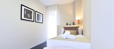 A hotel room with a bed, a chair, a desk with a lamp, and two framed pictures on the wall.