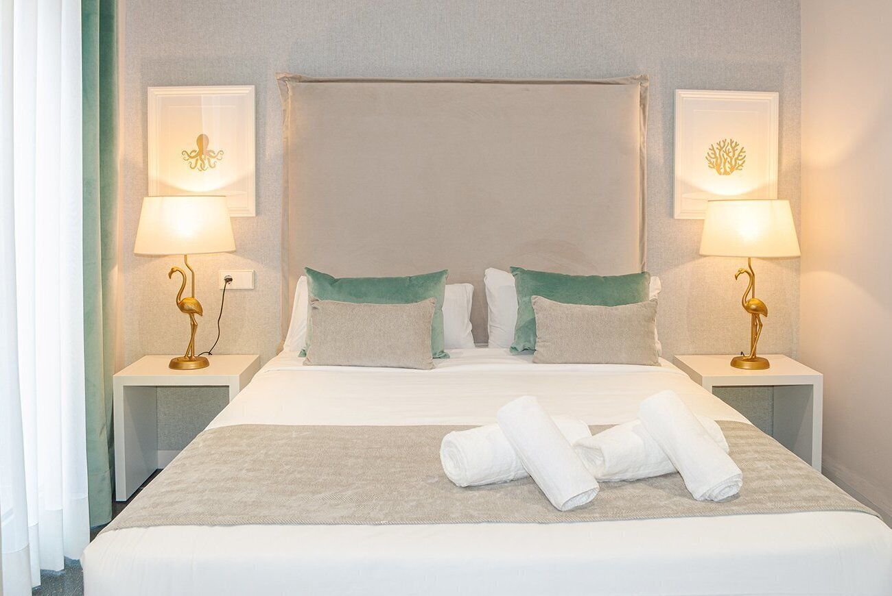 A neatly made bed with white linens and green pillows, two bedside lamps, and a pair of rolled towels on the bed.