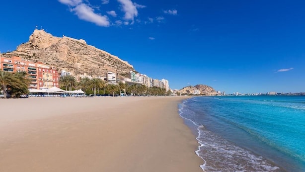 Una playa con una montaña al fondo, cielo azul despejado y edificios a lo largo de la costa.
