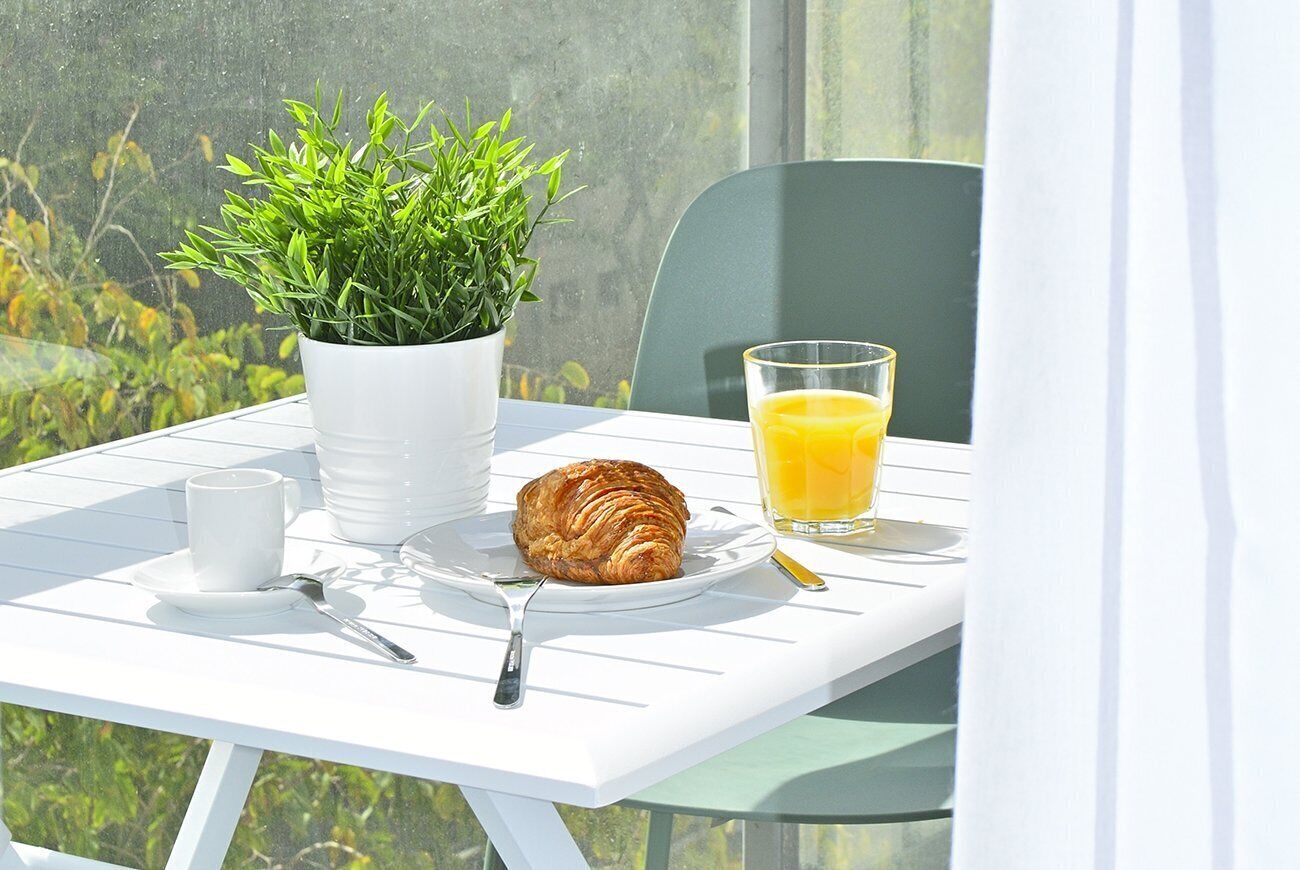 A breakfast setup on a white table with a potted plant, a croissant, a glass of orange juice, a cup, and cutlery.