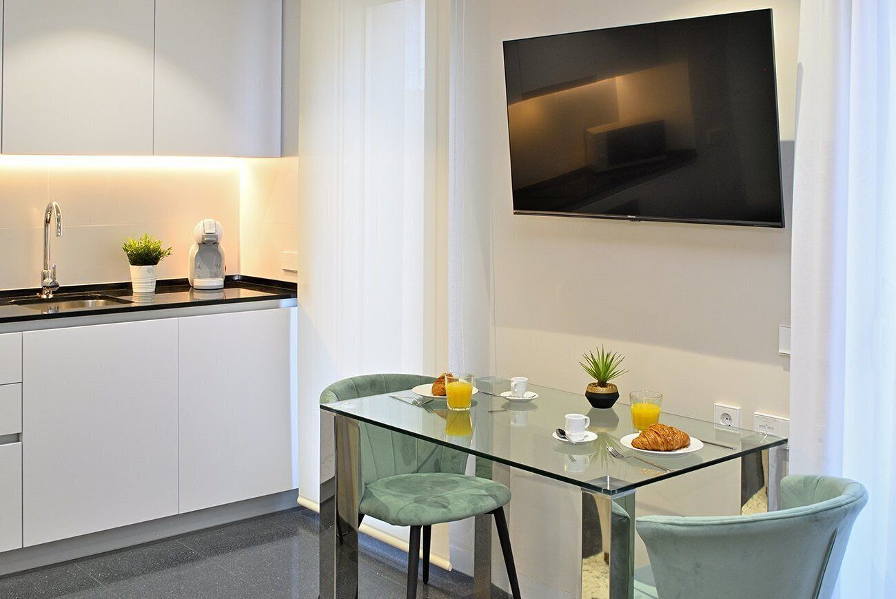 A modern kitchen with white cabinets, a black countertop, and a glass dining table with two chairs. A flat-screen TV is mounted on the wall above the table.