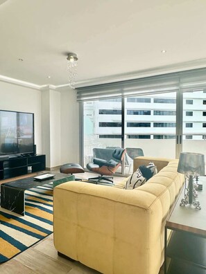 Un salon moderne avec un grand canapé, un téléviseur et une vue sur un immeuble à travers de grandes fenêtres.