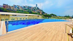 Una piscina con bordo in legno, un edificio con la scritta 'UNIDAD EDUCATIVA' e una collina con edifici residenziali sullo sfondo.