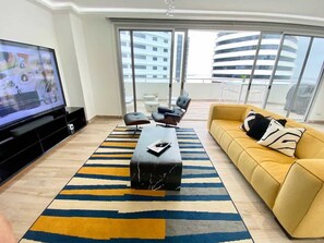 A modern living room with a flat-screen TV, a sofa, two armchairs, a coffee table, and a large window with a view of buildings.