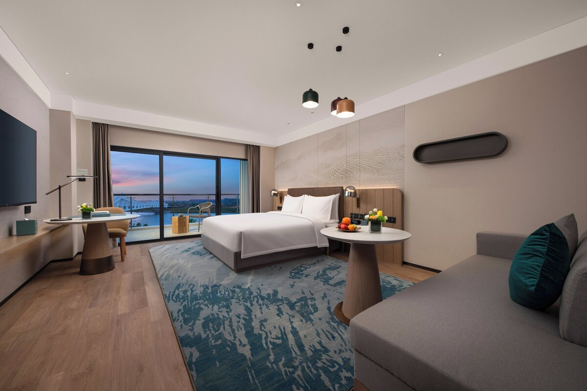 A modern hotel room with a large bed, a desk, a sofa, and a view of a bridge.