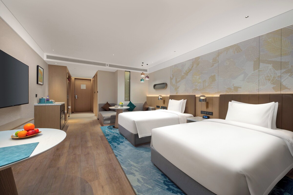 A hotel room with two beds, a desk with fruit, a TV, and a map on the wall.