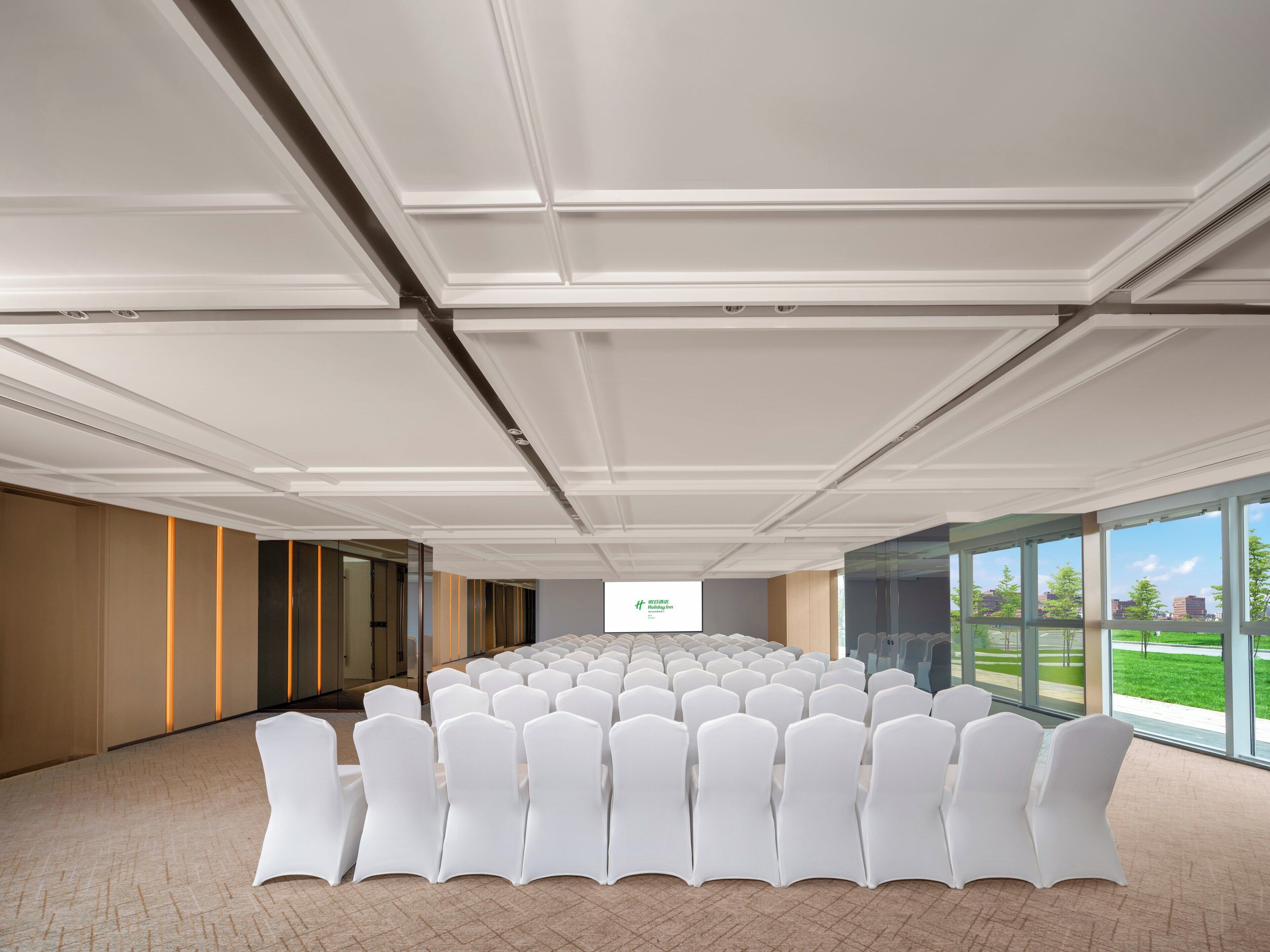 A conference room with rows of white chairs, a large screen at the front, and large windows offering a view of the outdoors.