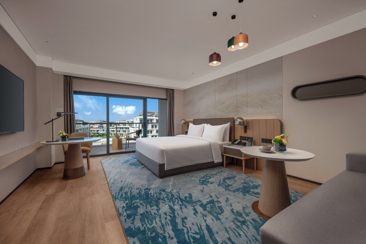A modern hotel room with a large bed, a desk, a chair, a round table with a vase of flowers, and a view of buildings through the window.