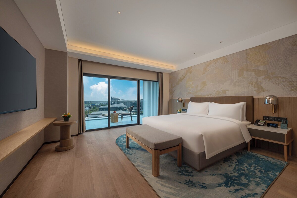 A modern hotel room with a large bed, a bedside table with a lamp, a small table with a plant, a bench, and a view of a cityscape through large windows.