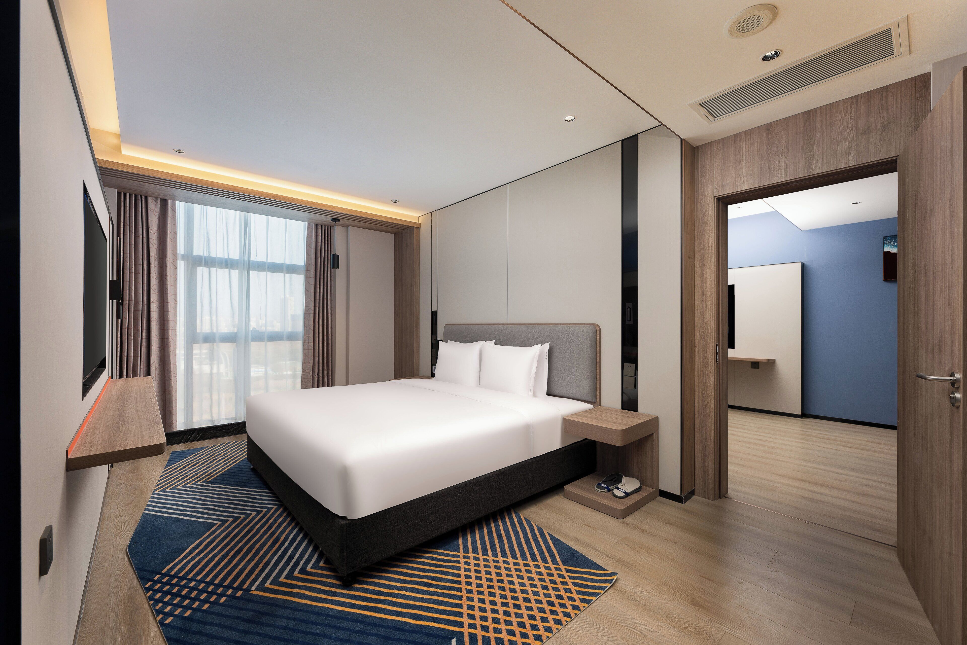 A modern hotel room with a large bed, bedside tables, a TV, and a view of the city through large windows.