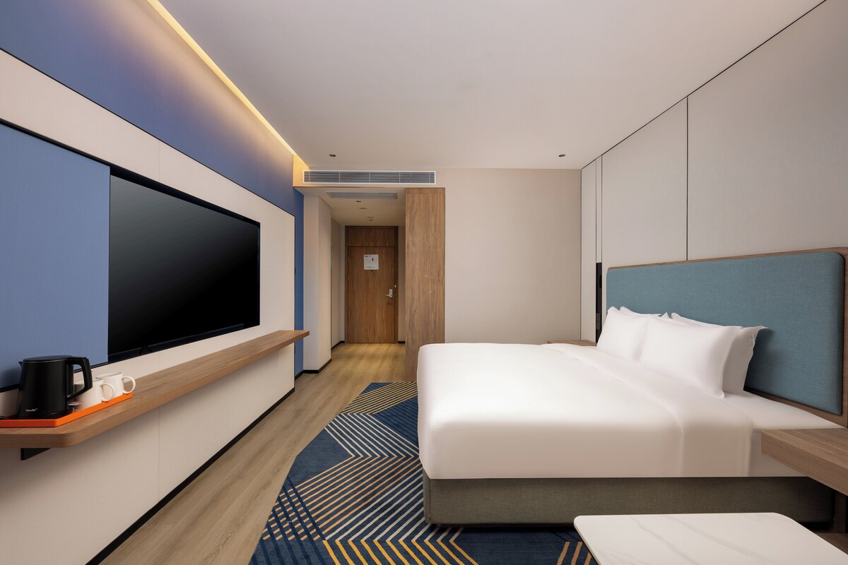 A modern hotel room with a large flat-screen TV, a bed with a blue headboard, a wooden nightstand, and a patterned rug.