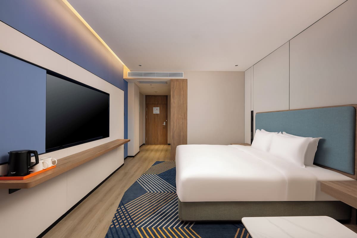 A modern hotel room with a large flat-screen TV, a bed with a blue headboard, a wooden nightstand, and a patterned rug.