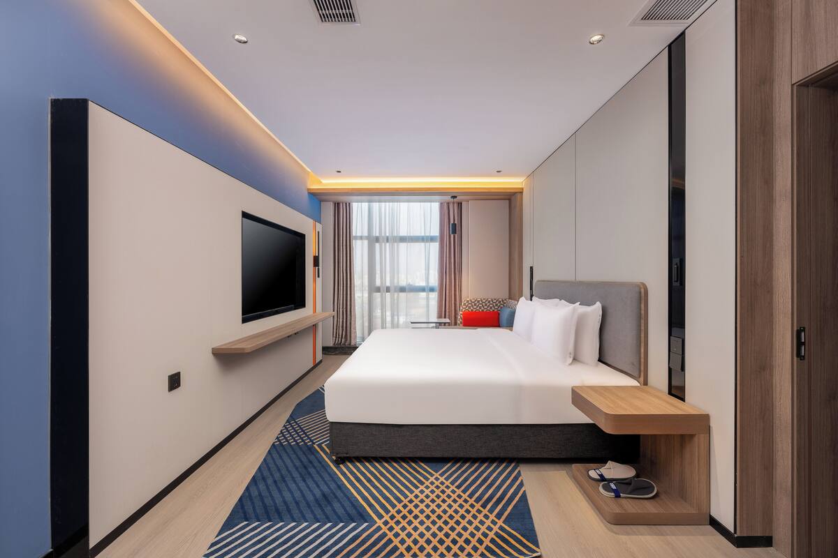 A modern hotel room with a large bed, a flat-screen TV mounted on the wall, a wooden bedside table, and a built-in bench.