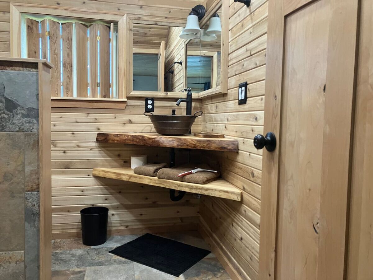 L’intérieur d’une cabane en bois, avec un mur en pierre, une étagère en bois où sont posées des serviettes, un seau noir et une porte dotée d’une poignée noire.