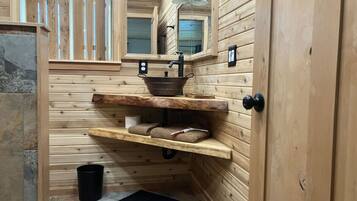 L’intérieur d’une cabane en bois, avec un mur en pierre, une étagère en bois où sont posées des serviettes, un seau noir et une porte dotée d’une poignée noire.