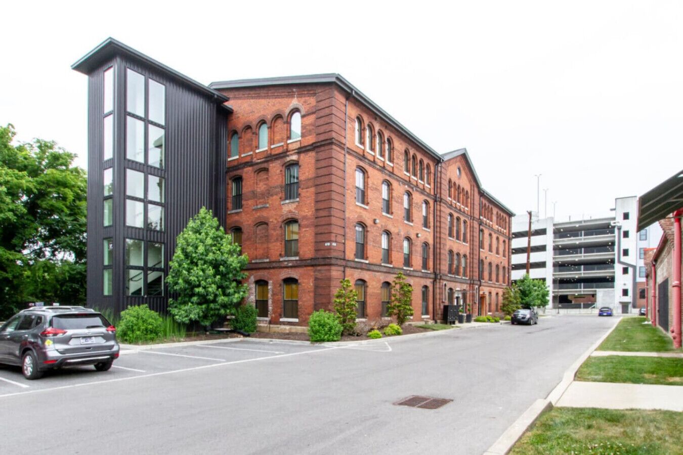 A street view with a modern building featuring large windows and a brick facade, adjacent to a parking garage.