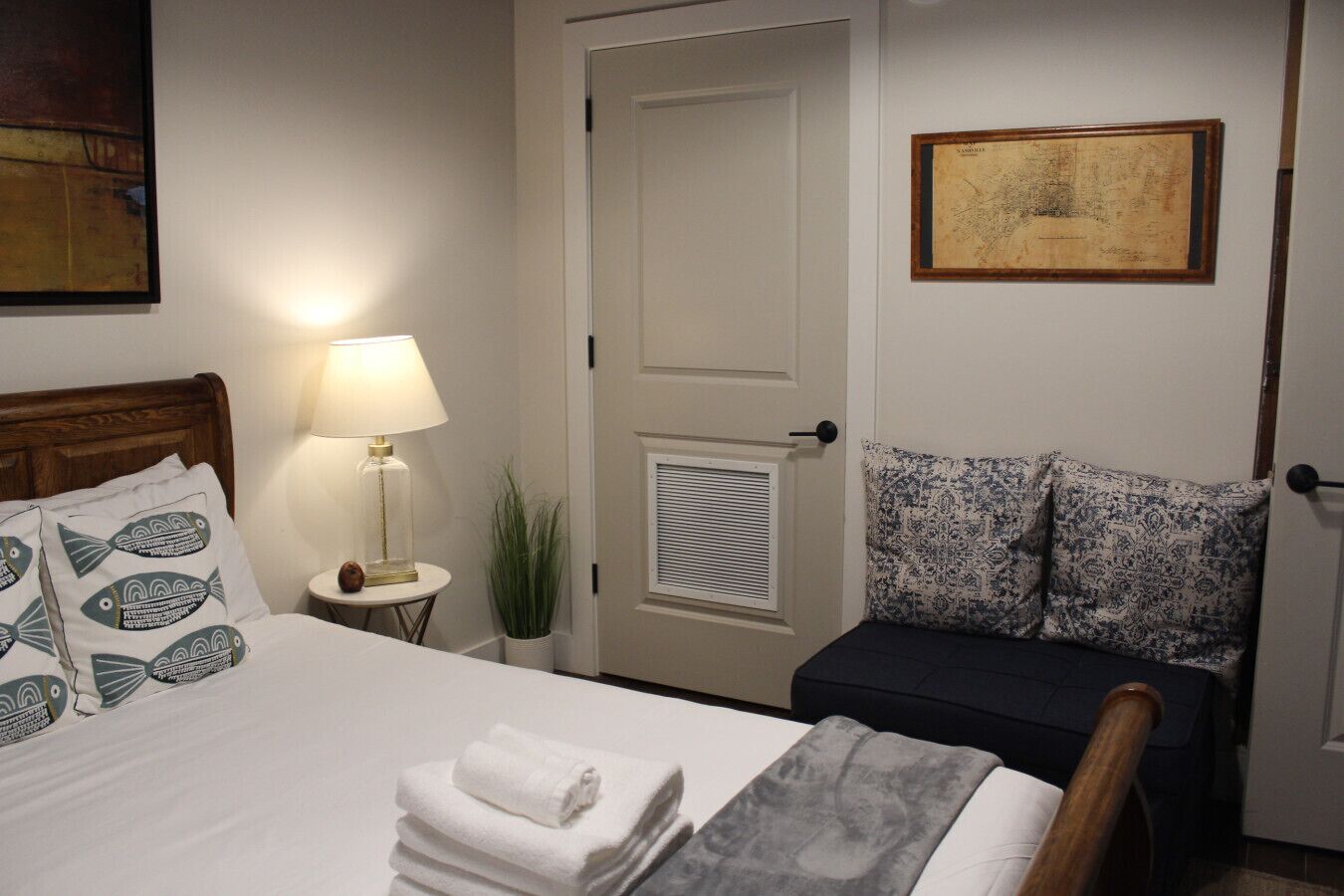 A neatly made bed with patterned pillows, a bedside lamp, a small plant, and a framed map on the wall.