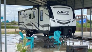 Zona de estacionamiento techada para vehículos recreativos, con una caravana, mobiliario exterior y una parrilla.