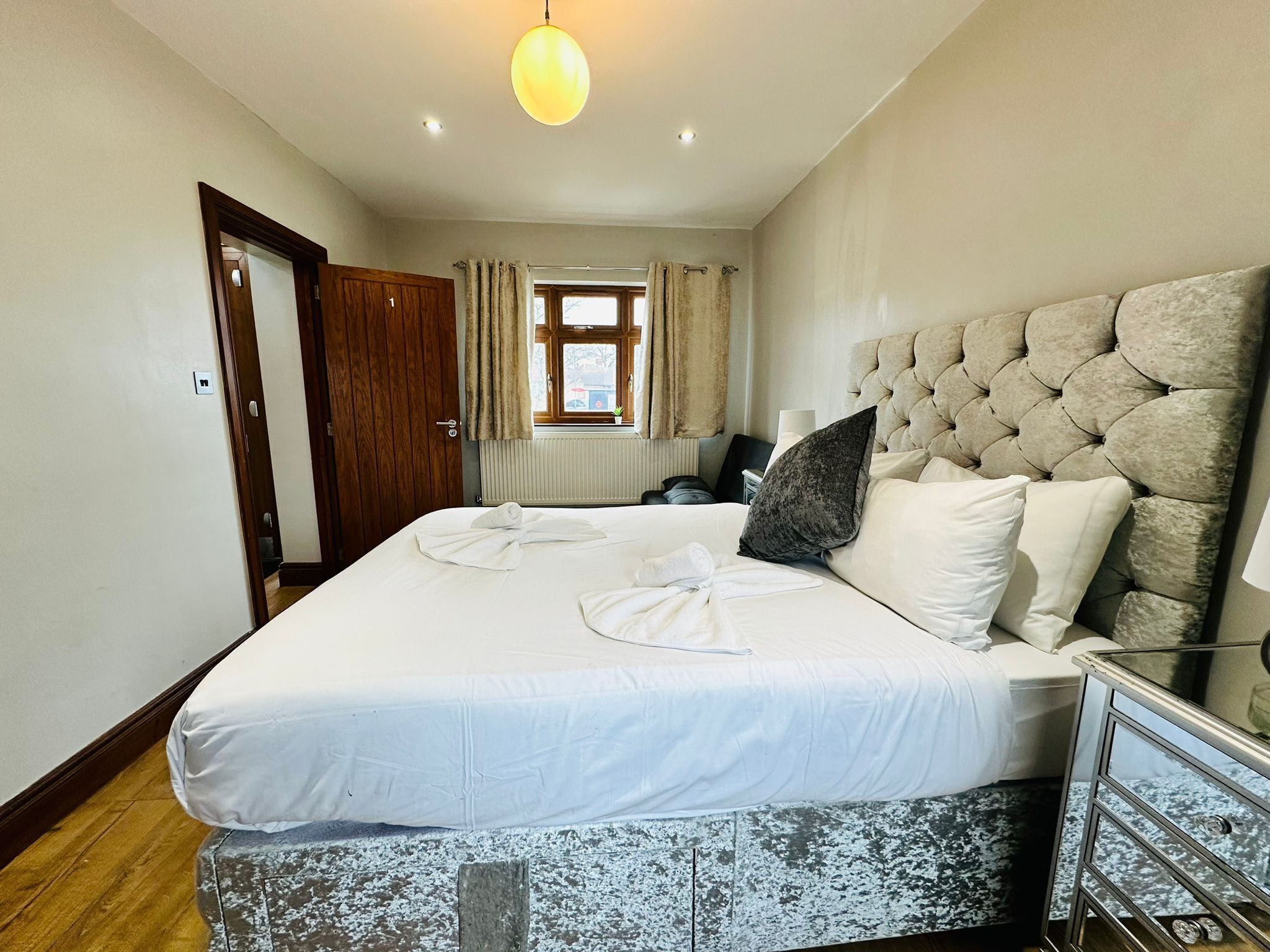 A hotel room with a large bed, a wooden door, a window with curtains, a radiator, and a bedside table with a lamp.