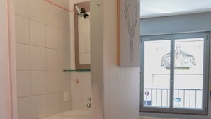 Une salle de bain avec un lavabo blanc, un miroir et des toilettes. Il y a une fenêtre donnant sur un balcon et un immeuble. Une petite table et un tapis sont posés sur le sol.