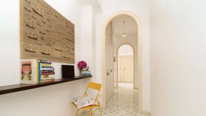 A hallway with a tiled floor, a wooden chair, a bookshelf with books and a laptop, and a wall with a maritime-themed artwork.