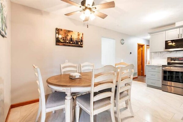 A dining area with a round table and white chairs, a ceiling fan, and a kitchen with modern appliances.