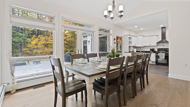 A dining area with a large table, chairs, and a view of autumn trees through large windows. - Luxurious Lake George Getaway (Lake George)