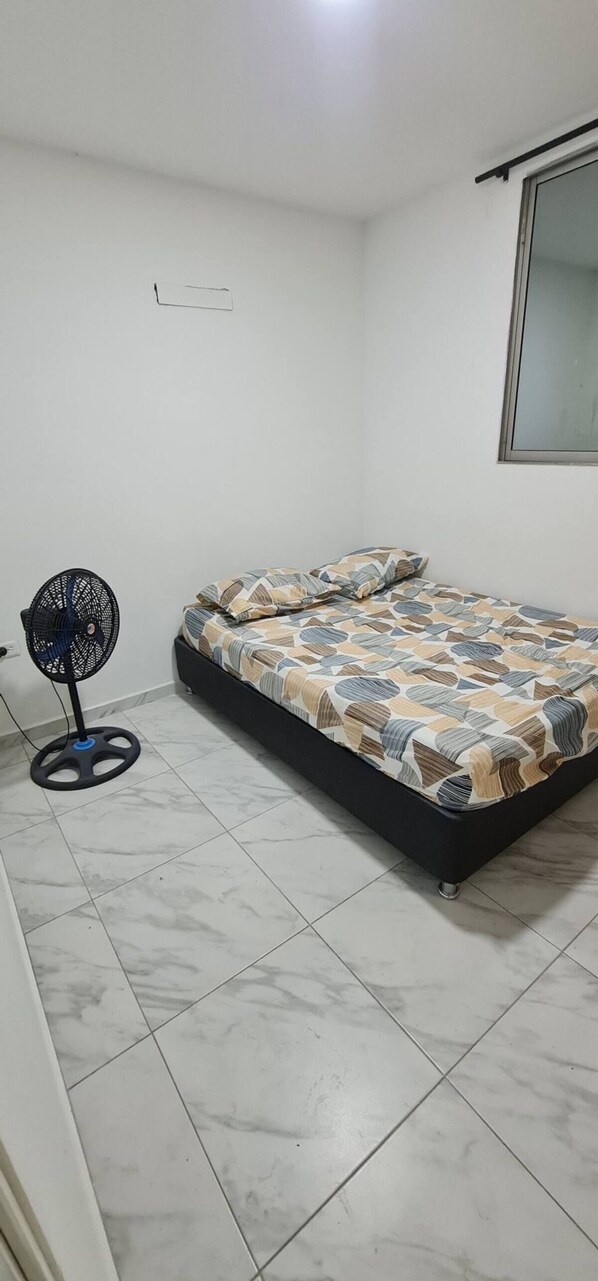 A bedroom with a bed, a standing fan, and a window.