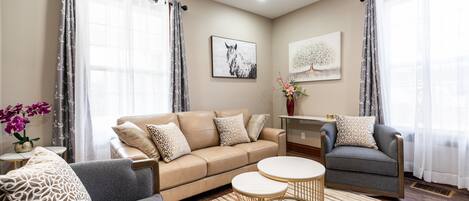 A living room with a beige sofa, two armchairs, a coffee table, and a rug. There are curtains, a painting, and a vase with flowers.