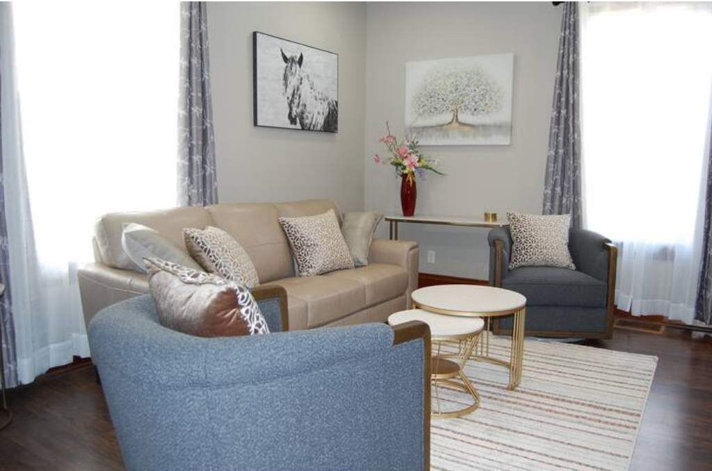 A living room with a beige sofa, a blue armchair, a round coffee table, and a grey armchair. There is a painting of a horse and a tree on the wall.