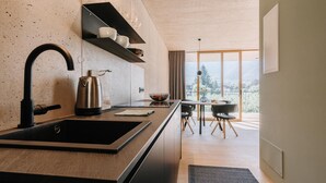 A modern kitchen with a large island, black faucet, and stainless steel kettle. There is a dining area with a table and chairs, and a view of trees through large windows. - Traumurlaub: Studio "apfel" auf dem Bio Bauernhof Langstrein (Laces)