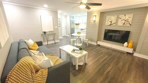 A modern living room with a gray sofa, a coffee table, and a TV. There is a dining area with a table and chairs. - FIFA 2026 Walk to Stadium | 1BR Unit 15L ATL (Atlanta)