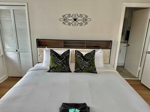 Un lit bien fait, agrĂ©mentĂ© de deux coussins dĂ©coratifs, avec une tĂȘte de lit en bois, une horloge murale et une porte fermĂ©e.