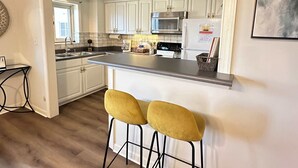 A modern kitchen with white cabinets, a stainless steel range hood, and a built-in microwave. There are two yellow bar stools and a small island with a sink. - FIFA 2026 Walk to Stadium | 1BR Unit 7-O ATL (Atlanta)