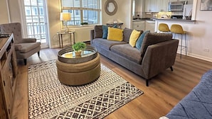 A living room with a grey sofa, a round coffee table with a plant, a patterned rug, and a kitchen in the background. - FIFA 2026 Walk to Stadium | 1BR Unit 7-O ATL (Atlanta)